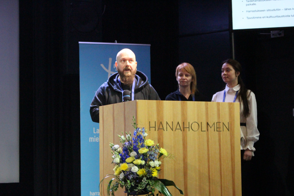 Marko Saarelainen puhuu puhujanpöntössä ja hänen vieressään seisovat Pauliina Lintula ja Sofiia Yakovyshyna.