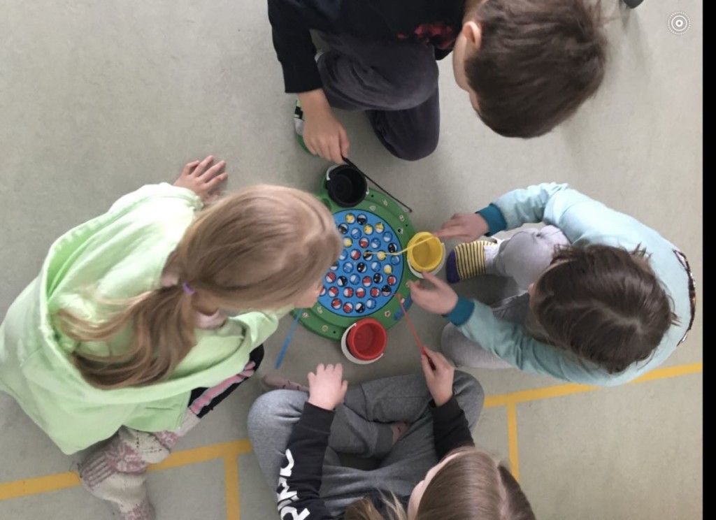 Neljä lasta istuu lattialla piirissä ja pelaa peliä, joka on heidän keskellään.
